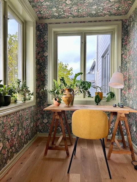 Et lite hjemmekontor med et trebord, en gul stol, vektorkirke, og flere planter, omgitt av floral tapet og store vinduer som slipper inn naturlig lys.