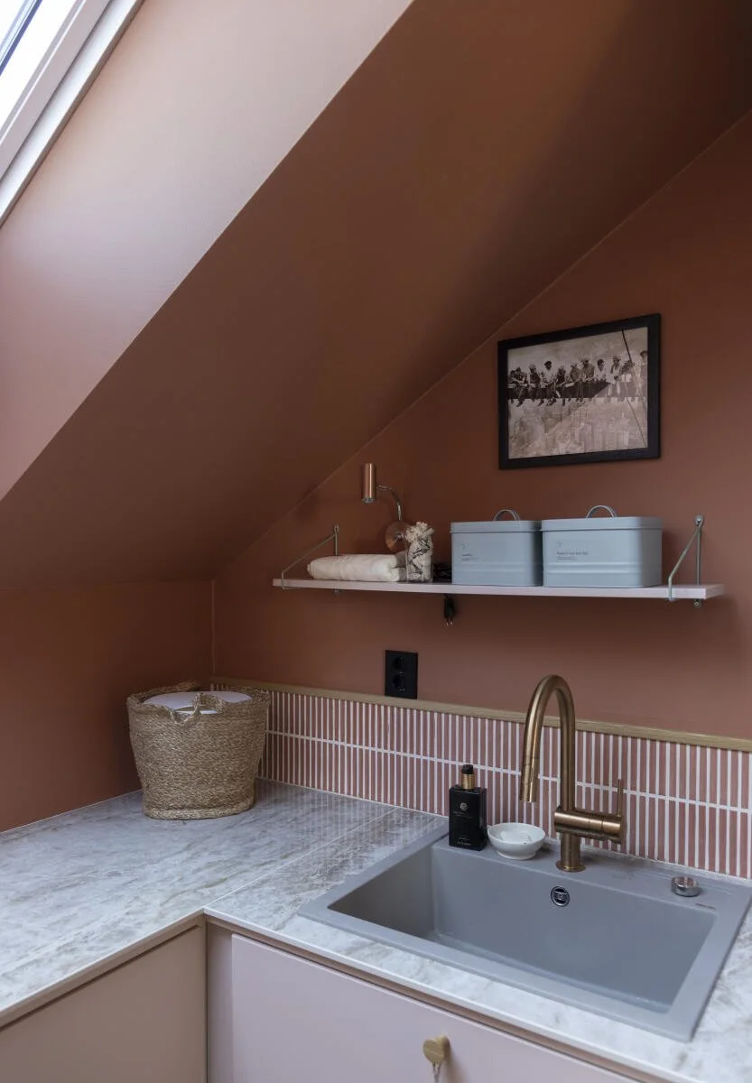 Kjøkkenvask med en kran, soppskål og ulike oppbevaringsbokser. Veggdekorasjon med et svart-hvitt bilde og en liten hylle med håndklær og dekorasjoner. Rektangulært vindu og en kurv på benkeplaten. Foto: Filippa Tredal