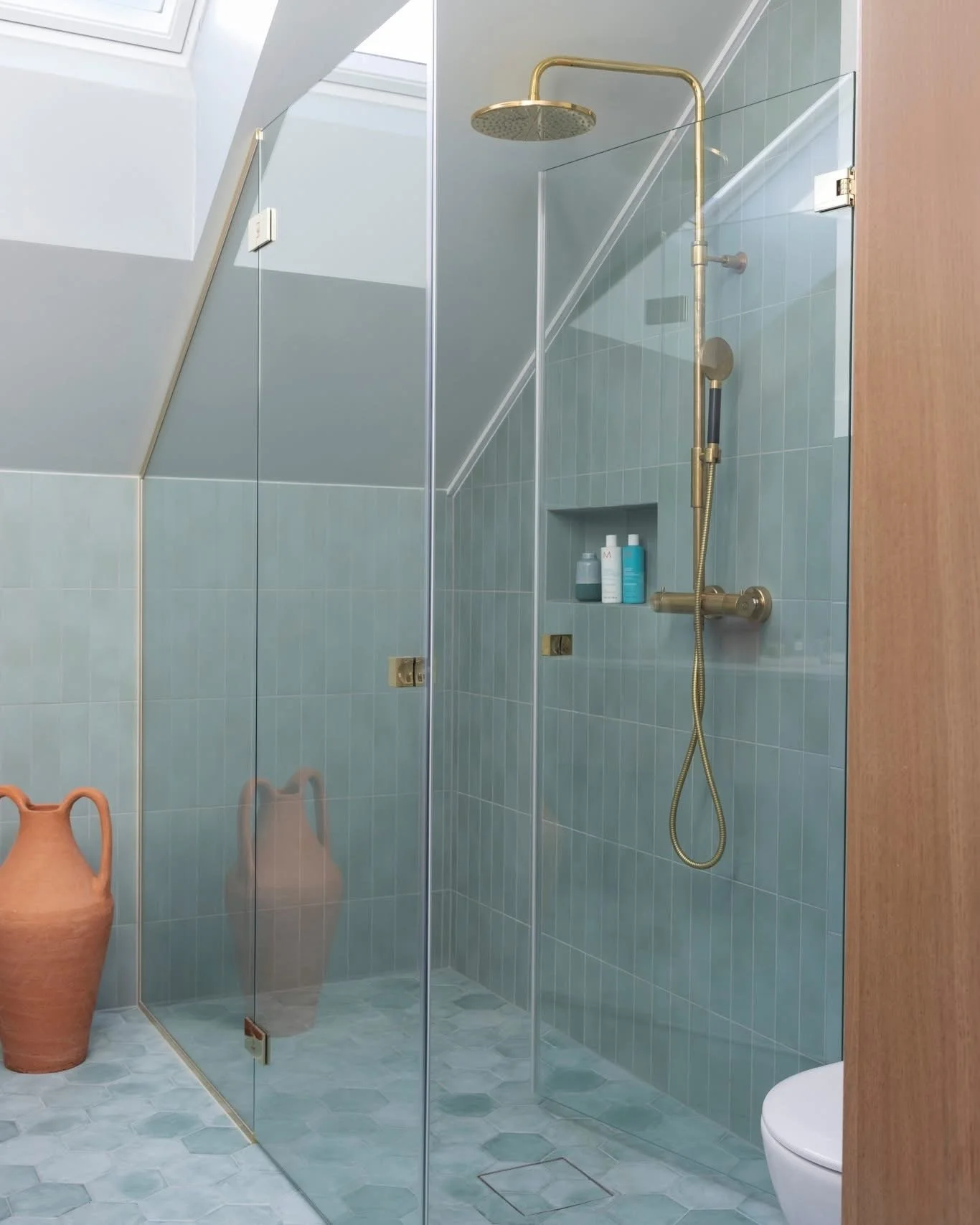 Et bad med en glassvegg, dusjhode og blå fliser på veggene. Det er også to terrakottakanner på gulvet og en badekar i nærheten. Foto: Filippa Tredal