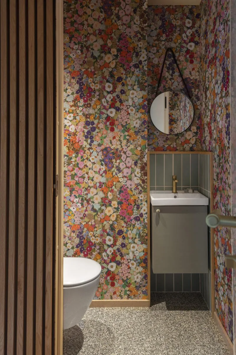 Liten bad med veggdekor med blomstermønster, speil over liten vask, toalett til venstre, og tre paneler av tre på venstre side.Foto: Filippa Tredal