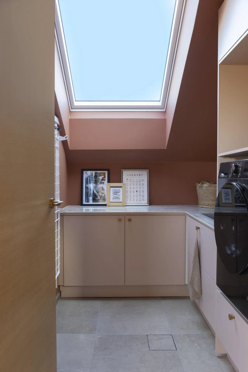 Et vaskerom med skråtak og takvindu, hvite skap og hyller, dekorative bilder og en kurv på benkeplaten, samt deler av en vaskemaskin på høyre side. Foto: Filippa Tredal