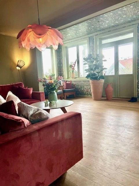 Stue med rosa sofa, blomster i grønne vaser, store planter, store vinduer, og et taklysdeksel som ligner en blomsterlignende lampe.