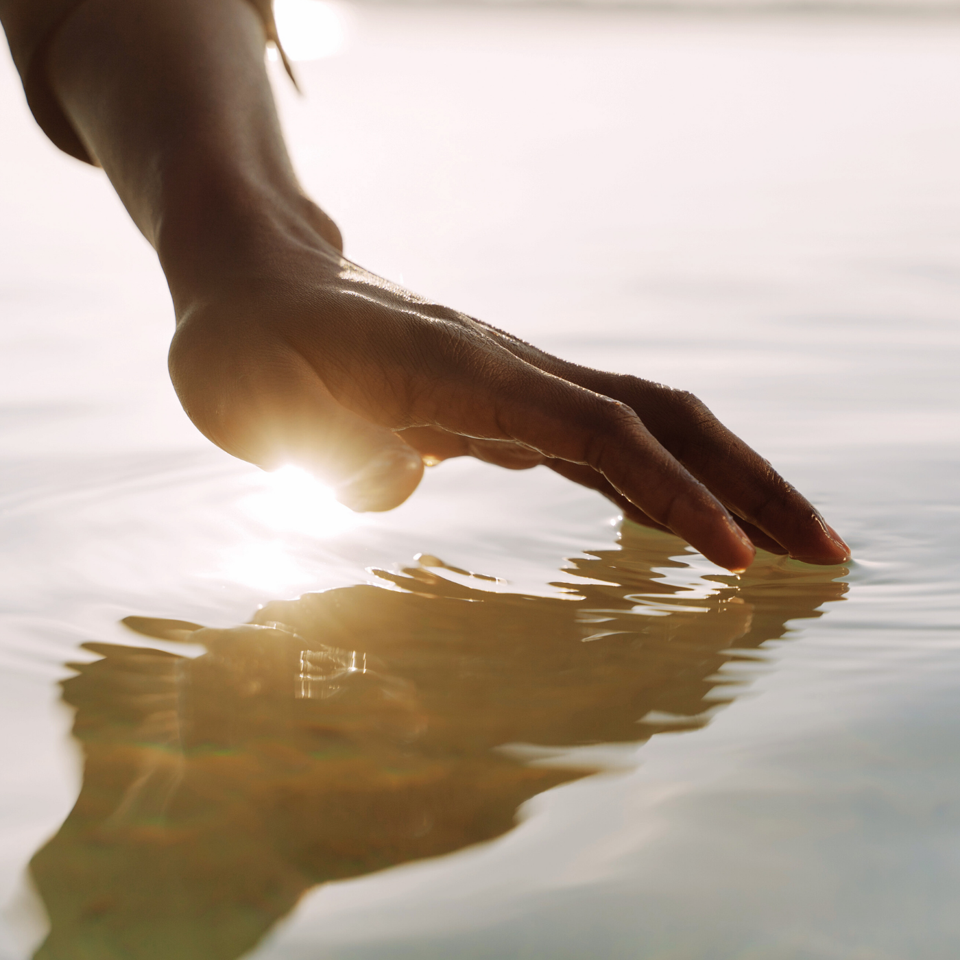 A person's hand reaches into calm water with sunlight reflecting off the surface.
