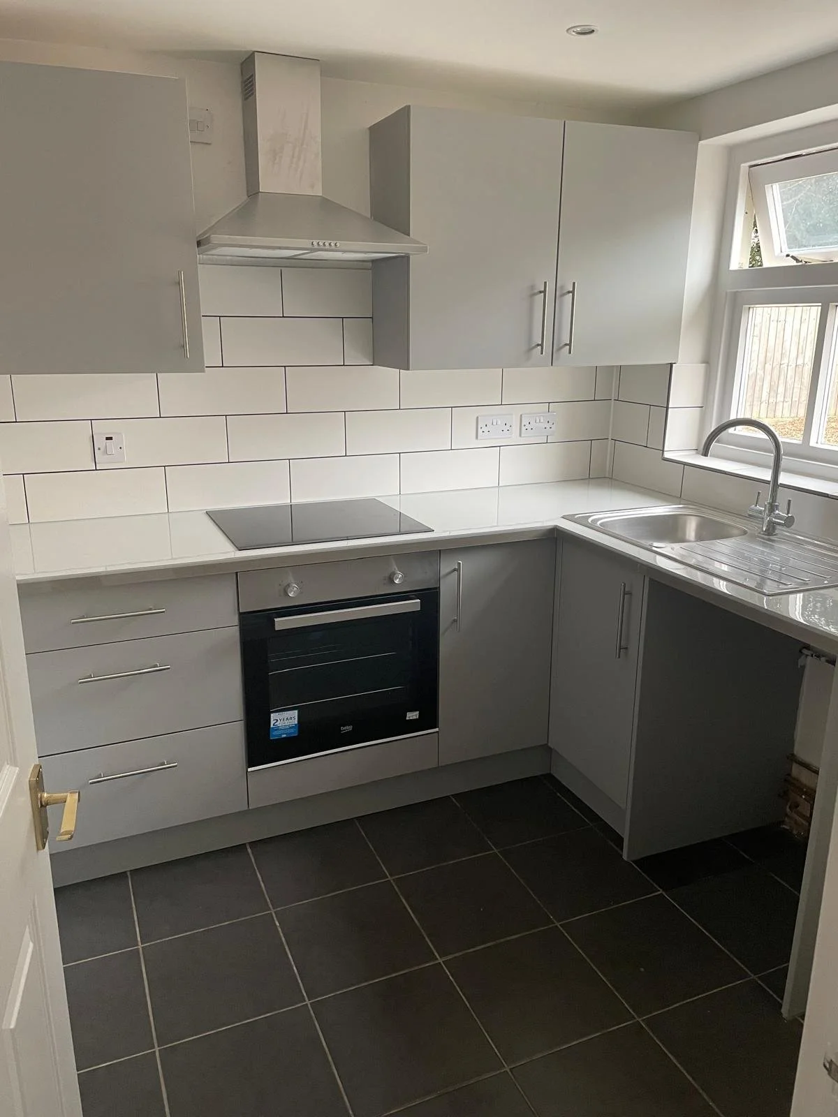 A modern kitchen with gray cabinets, white tiled backsplash, black oven, electric stove, stainless steel sink, and a window above the sink.