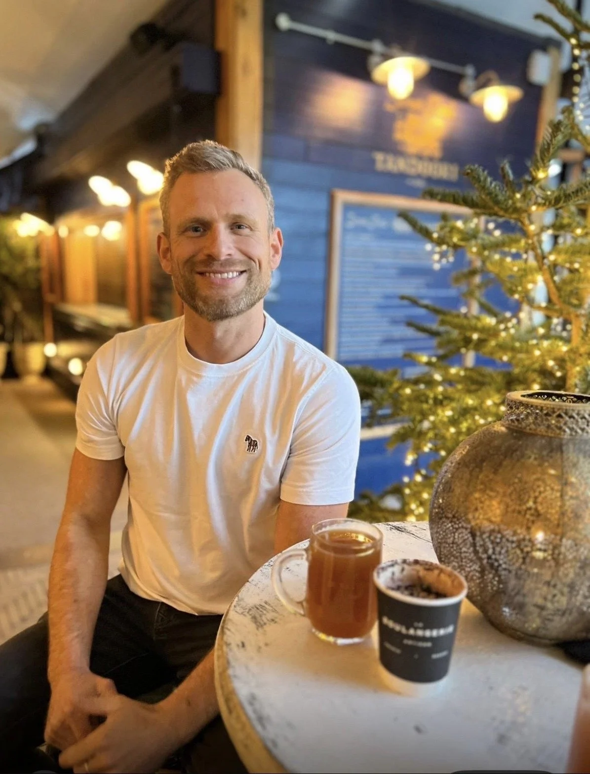 A smiling man with short hair and a beard sitting at a table with drinks, in a cozy, decorated restaurant or cafe with a Christmas tree and warm lighting.