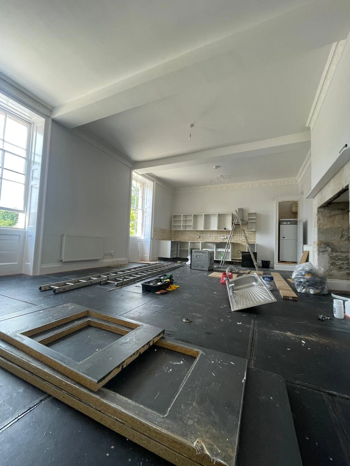 Living room under renovation with tools, materials, and construction debris scattered on the floor, and an open kitchen area in the background.