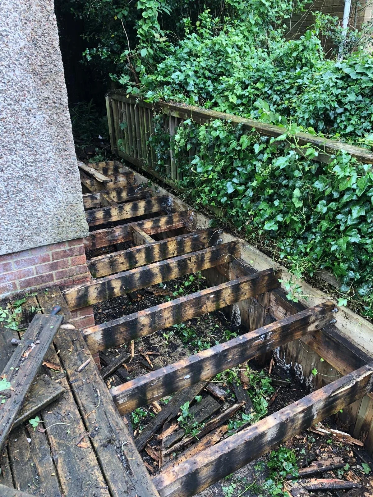 Partially demolished wooden deck with burned wood, next to a brick and stucco house, surrounded by dense green bushes.