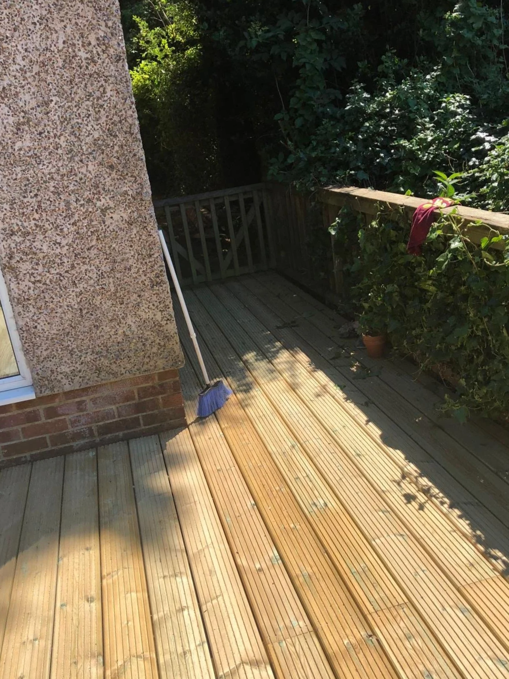 Sunlit wooden deck with a broom leaning against the house wall, a flowerpot, and green foliage surrounding the area.