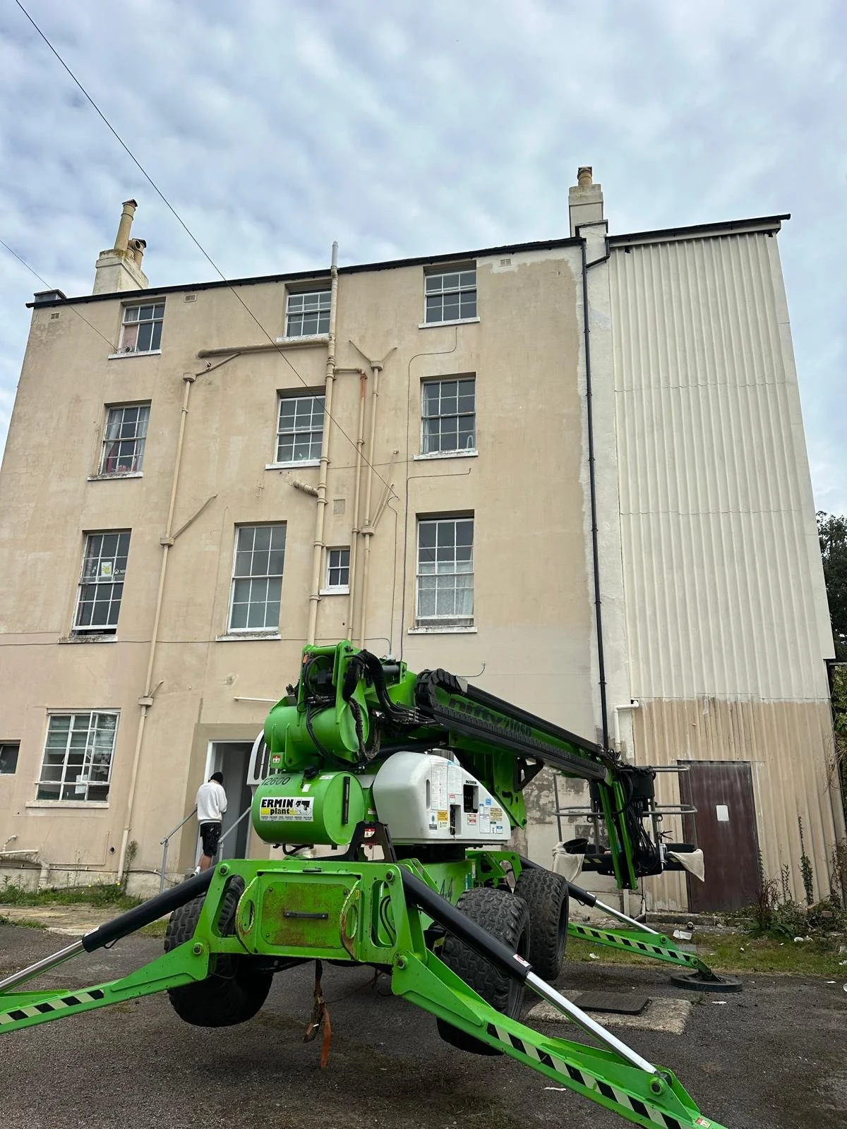 A green of the ground crane positioned in front of a beige residential building with multiple windows, under a cloudy sky.