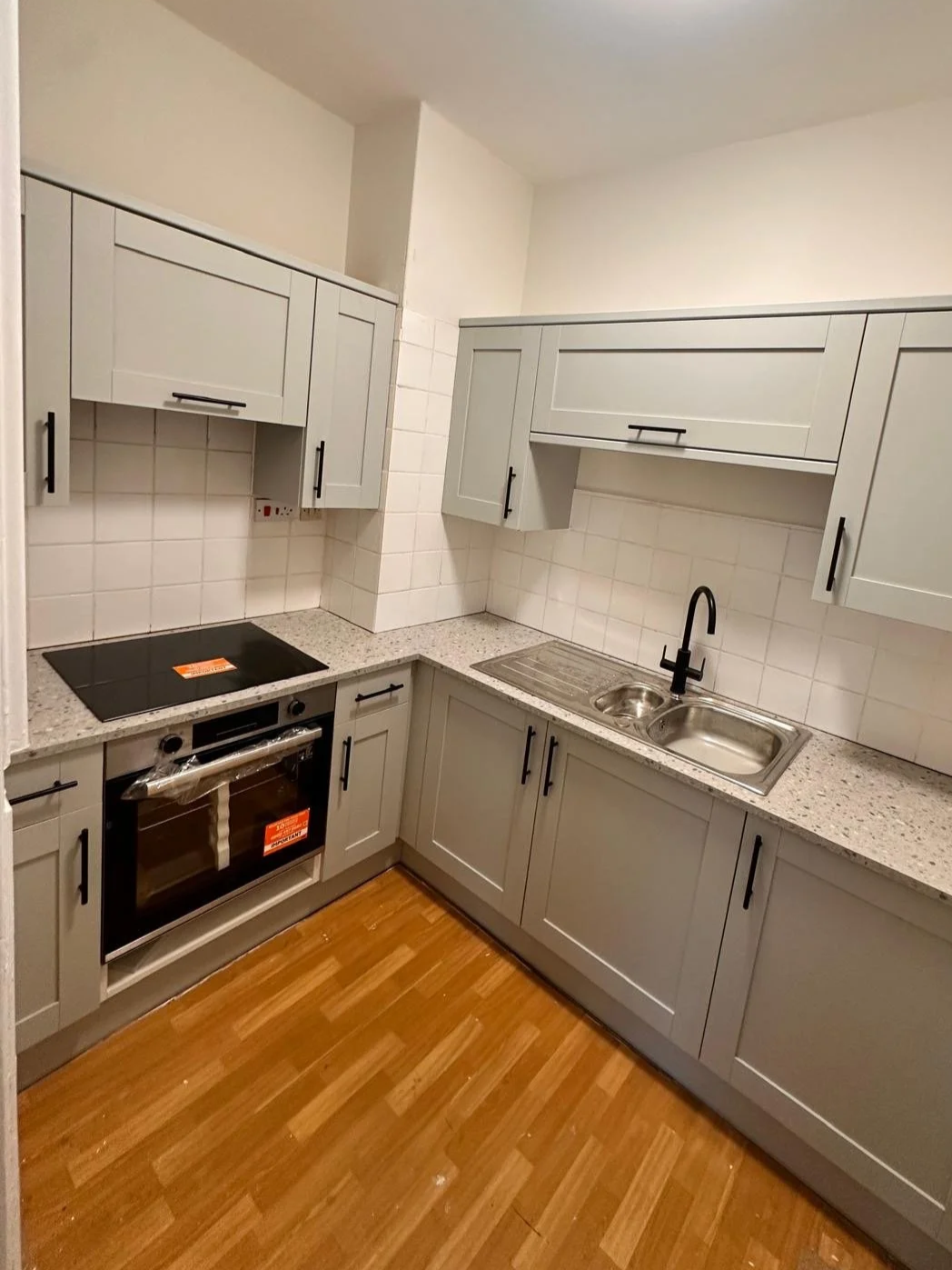 Small modern kitchen with gray cabinets, a black stovetop, an oven, a double sink with a black faucet, and wood flooring.