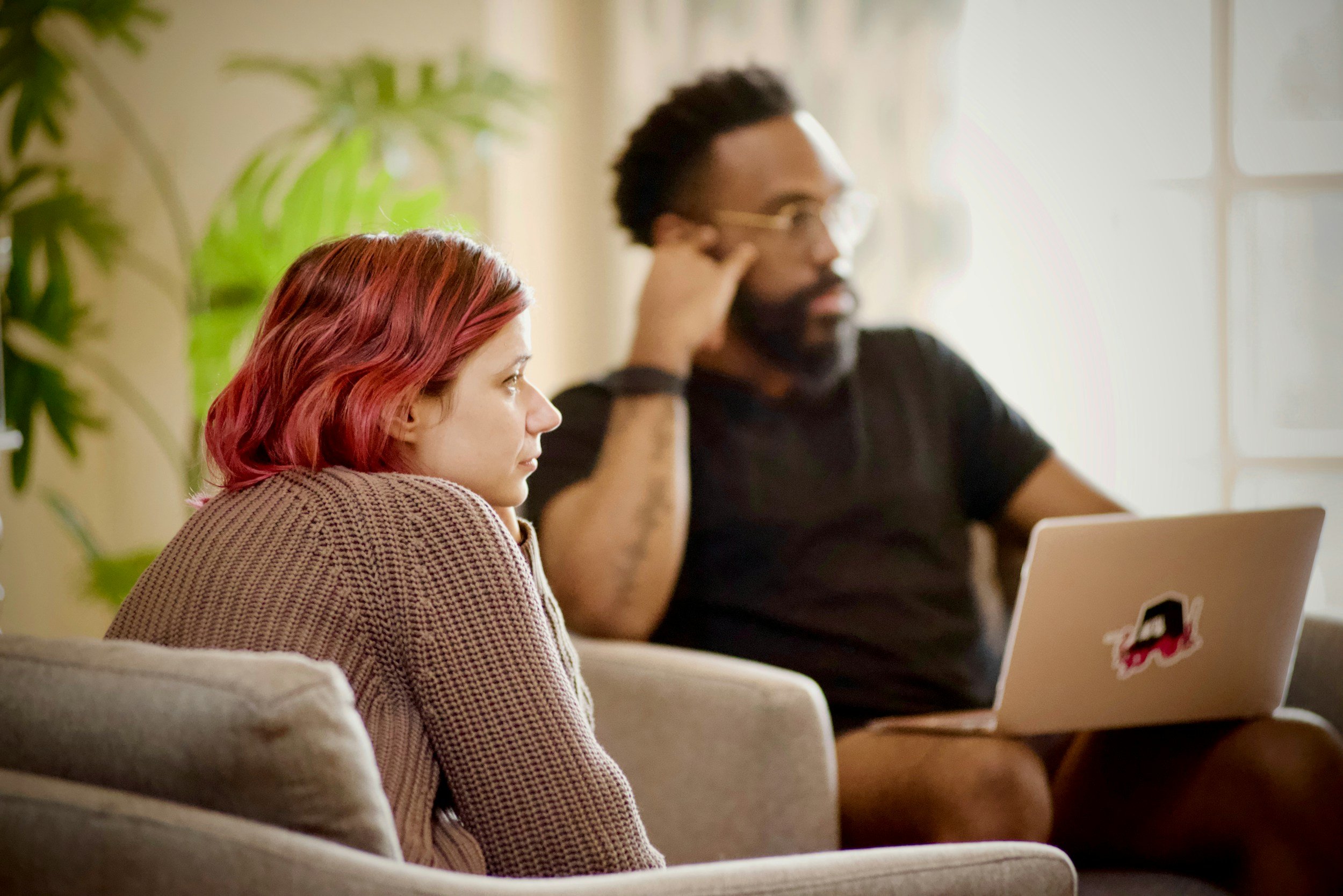 A woman with red hair and a man with glasses and a beard sitting on a beige couch in a bright room with large windows and a green plant.