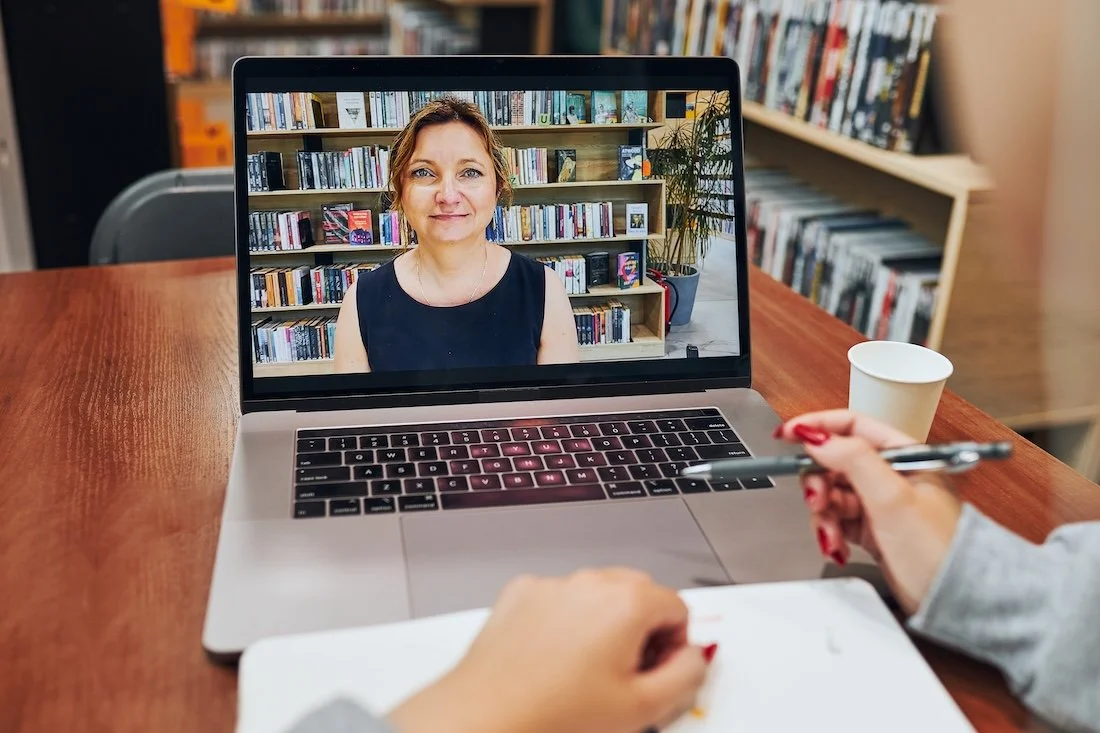 A person having a video call on a laptop with a woman in a library background. The person holds a pen and has a notepad in front. There is a paper cup next to the laptop.