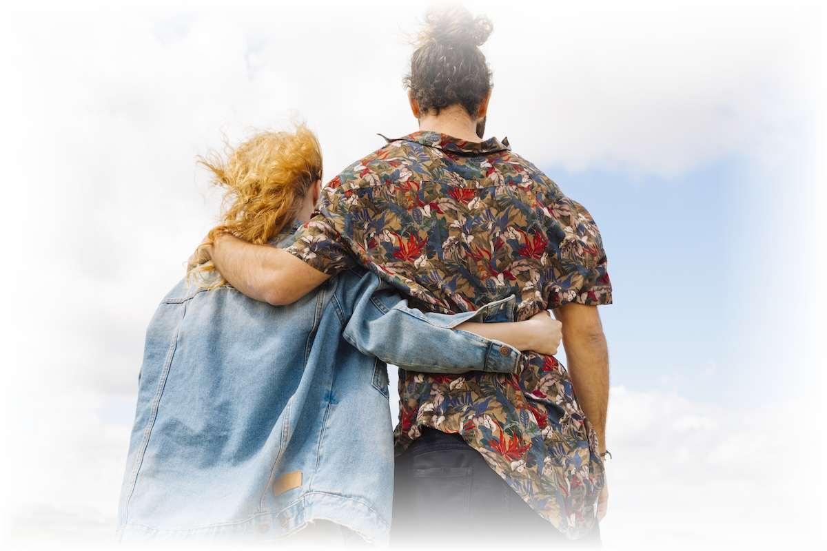 A person with curly red hair in a denim jacket and another person with curly brown hair in a floral shirt are embracing outdoors, facing away from the camera, under a partly cloudy sky.