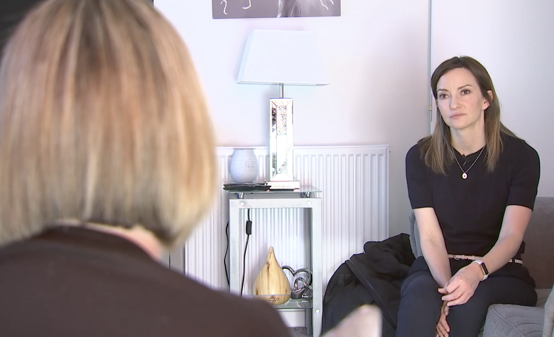 A woman with brown hair wearing a black shirt sitting in a room with white walls, a modern table lamp, and decorative objects including a white vase and a wooden ornament on a table behind her, during an interview or conversation.
