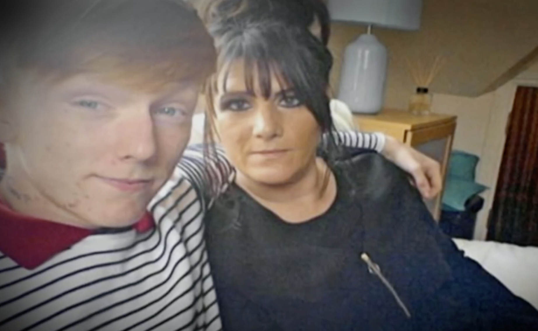 A man and woman taking a selfie indoors, with a lamp, table, and other furniture visible in the background.