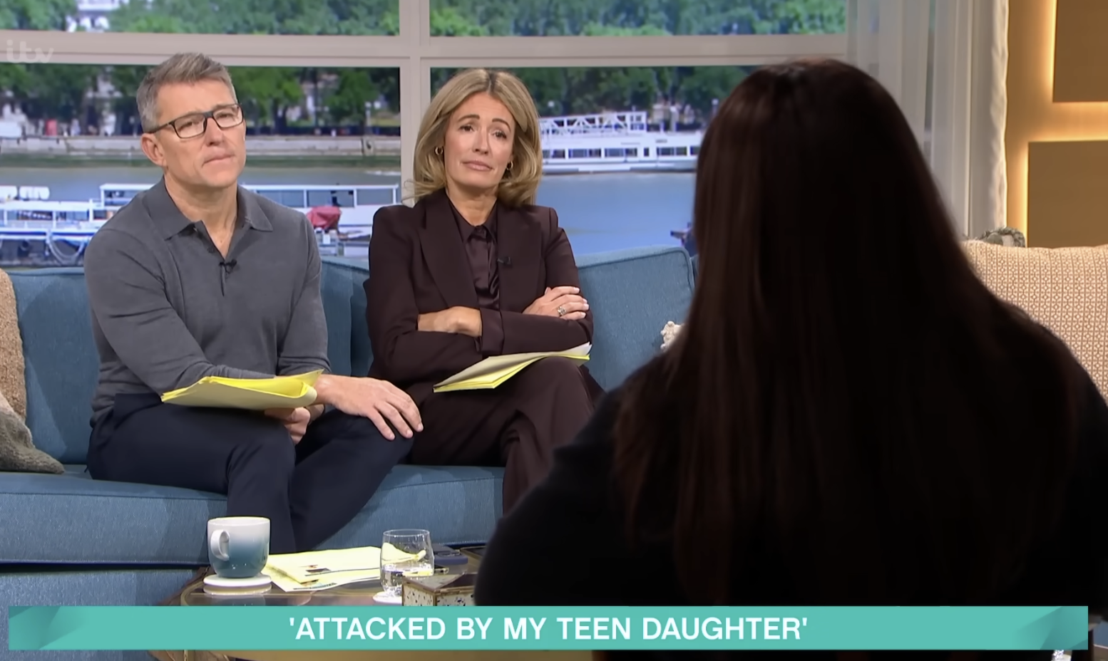 Television talk show with two hosts, a man and a woman, sitting on a blue couch and listening to a guest woman with long dark hair. The setting includes a window with a river view, mugs, glasses, papers, and microphones, with a text banner at the bottom reading 'Attacked by my teen daughter.'