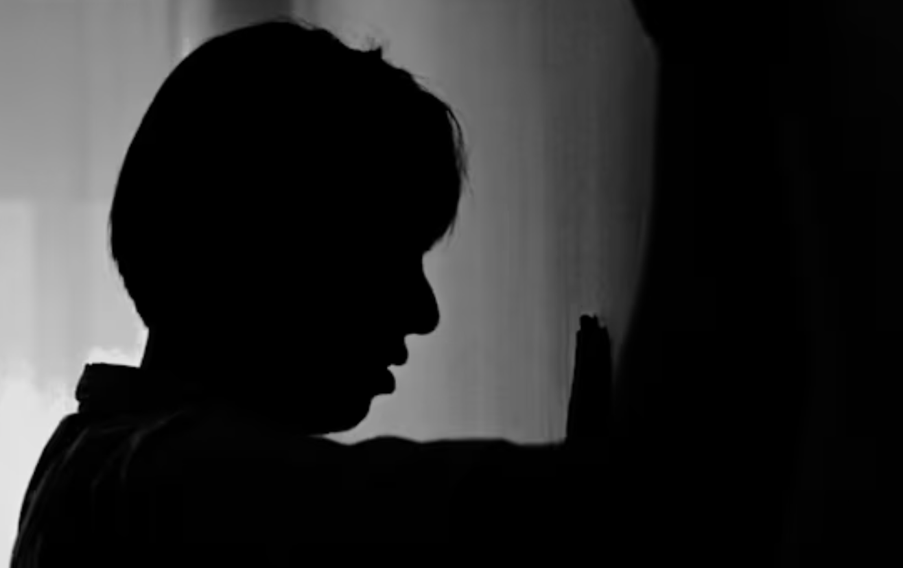 Silhouette of a young boy with short hair pressing his hand against a wall or door, in a black and white photo.