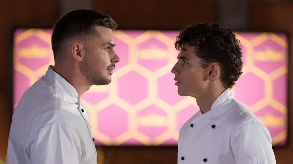Two male chefs in white uniforms facing each other in a kitchen with a pink and yellow patterned background.