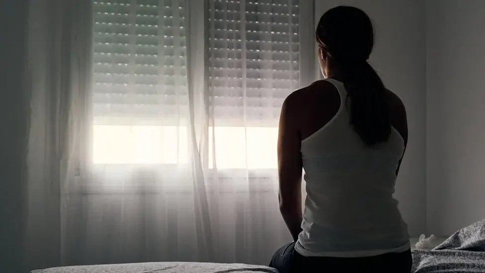 A person with dark hair in a ponytail, wearing a white sleeveless top, sitting on a bed and looking out a window with closed blinds and sheer curtains.