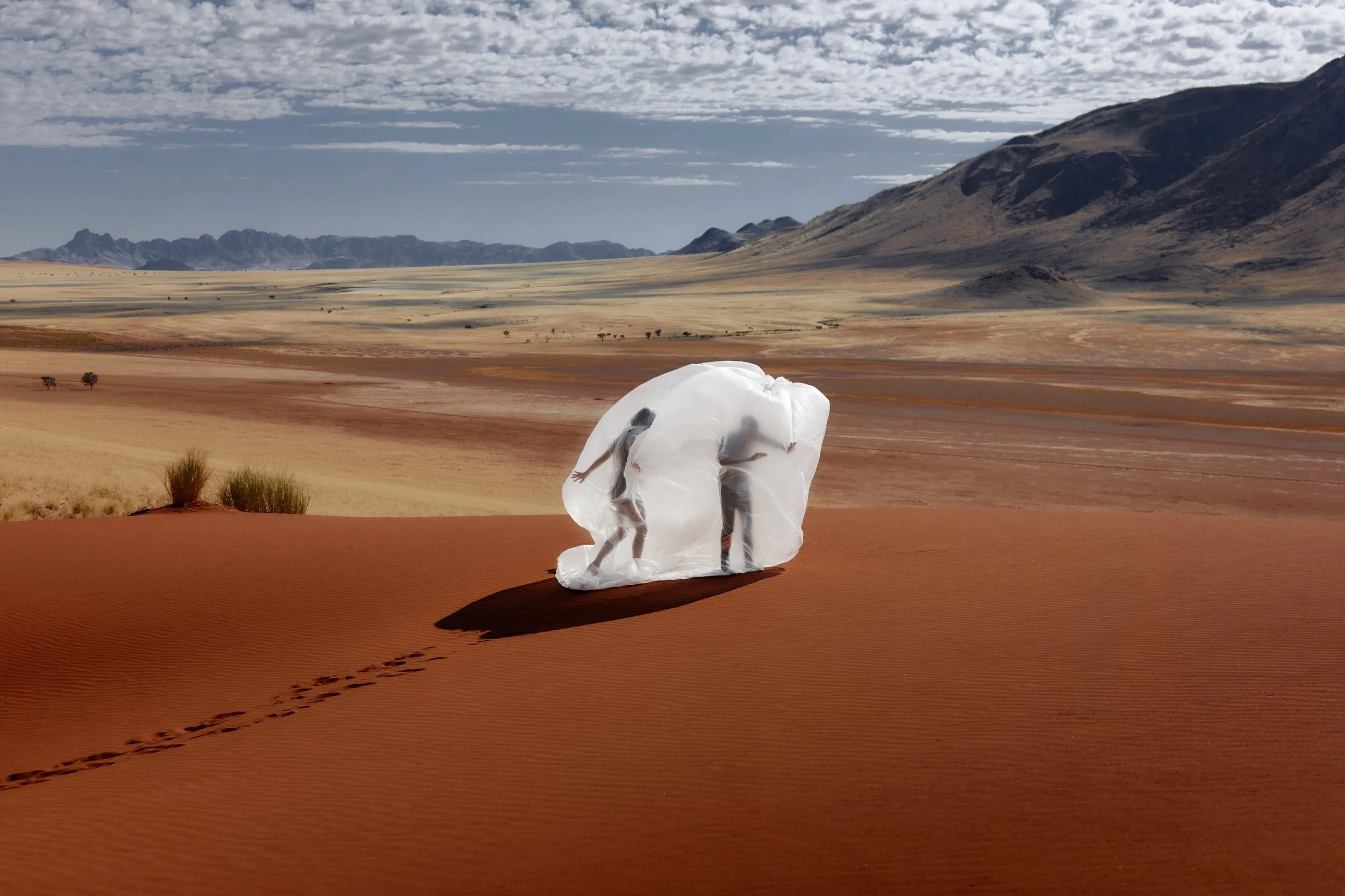 Two people wearing protective suits and masks walking through a desert landscape with mountains in the background.