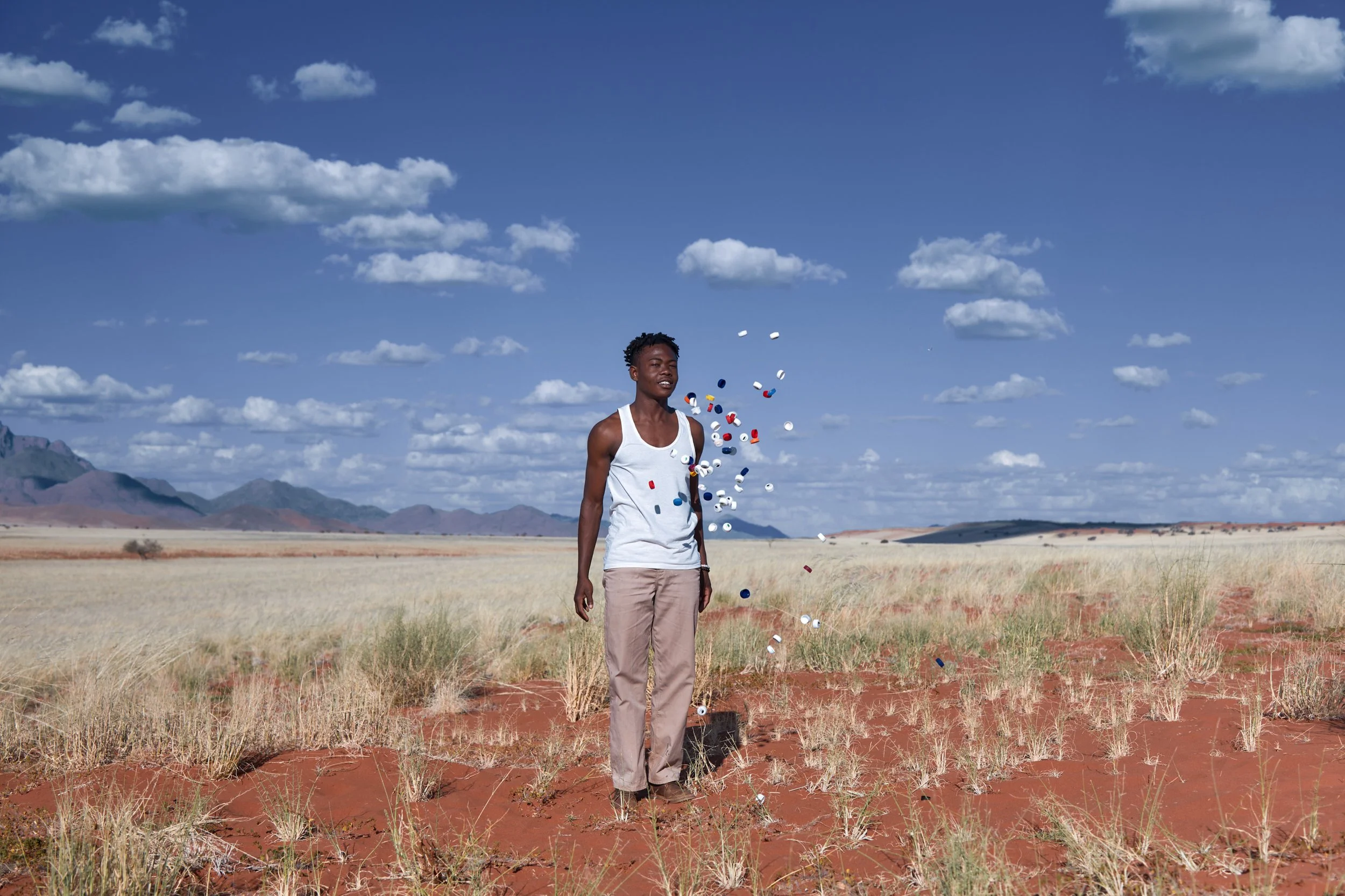 A young man standing in a vast open field with mountains in the distance, under a blue sky with scattered clouds, as multicolored pills or capsules are falling around him.