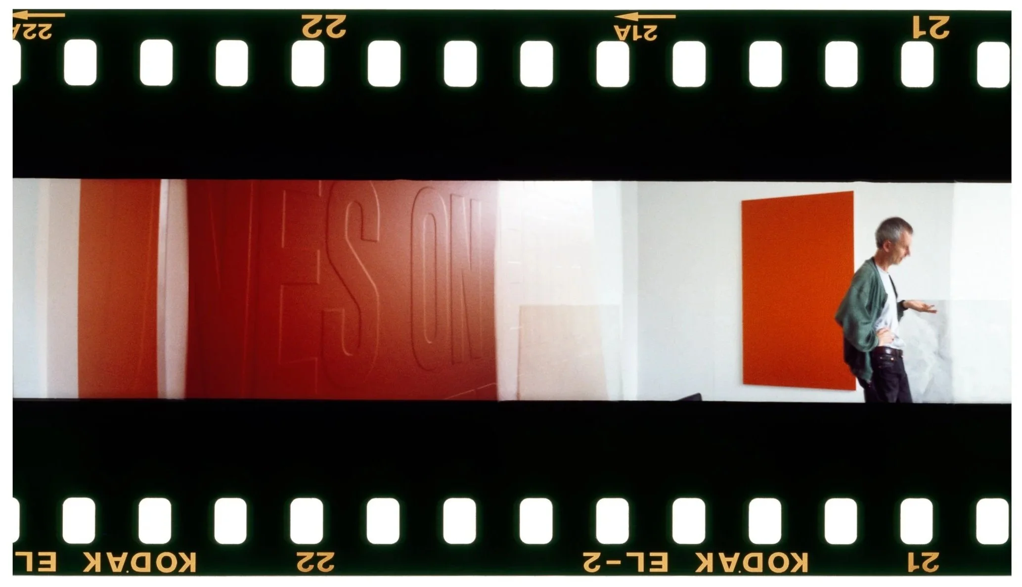 A person standing in a modern interior with red and white walls, one of which has a large embossed 'RES' and the other an orange rectangular panel, seen through a film strip border.