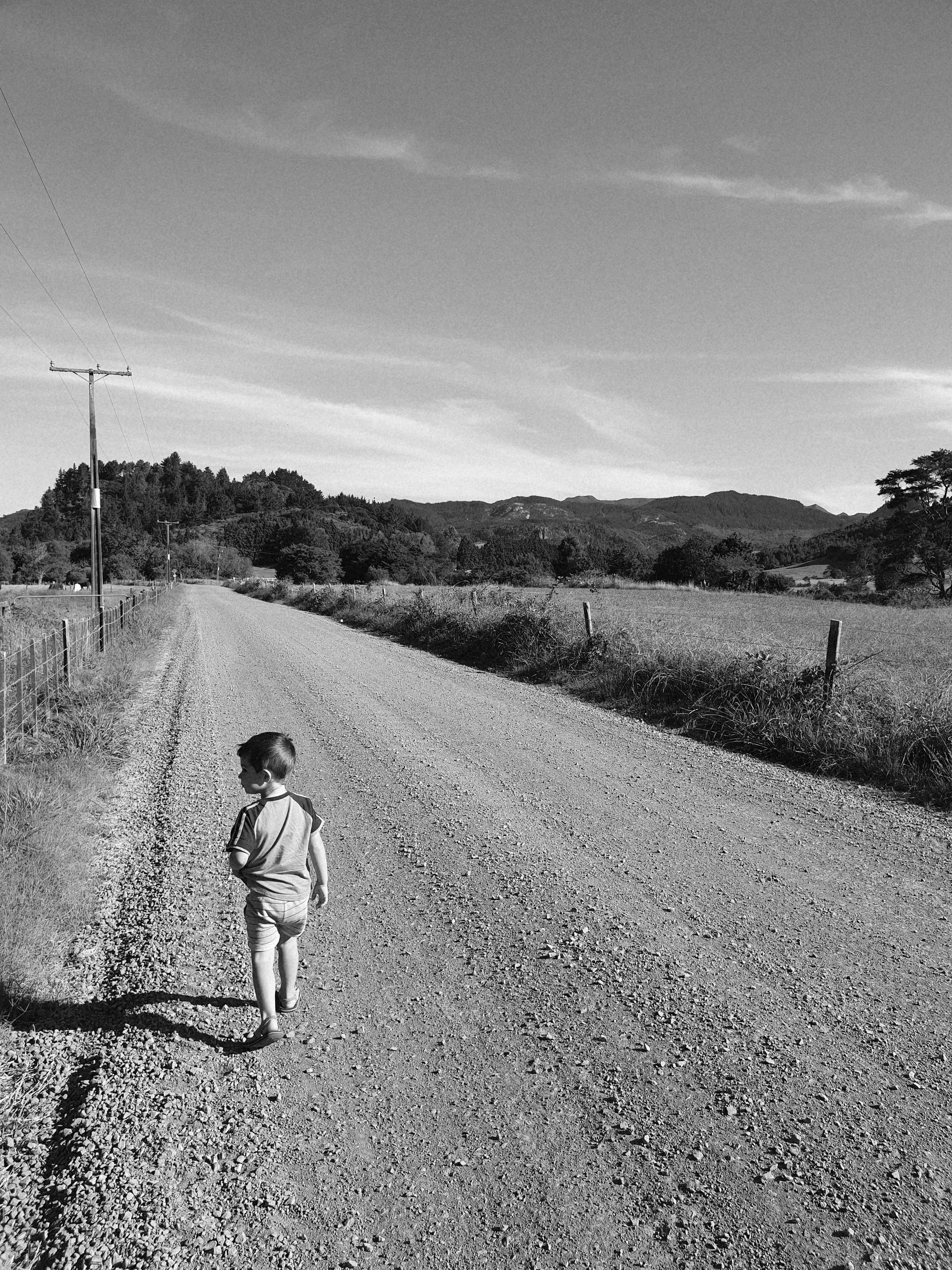 A black and white photo of a boy walking on a gravel road in a rural landscape. The scene features mountains in the background, a fence on the left side, and utility poles along the road. The sky shows some clouds.