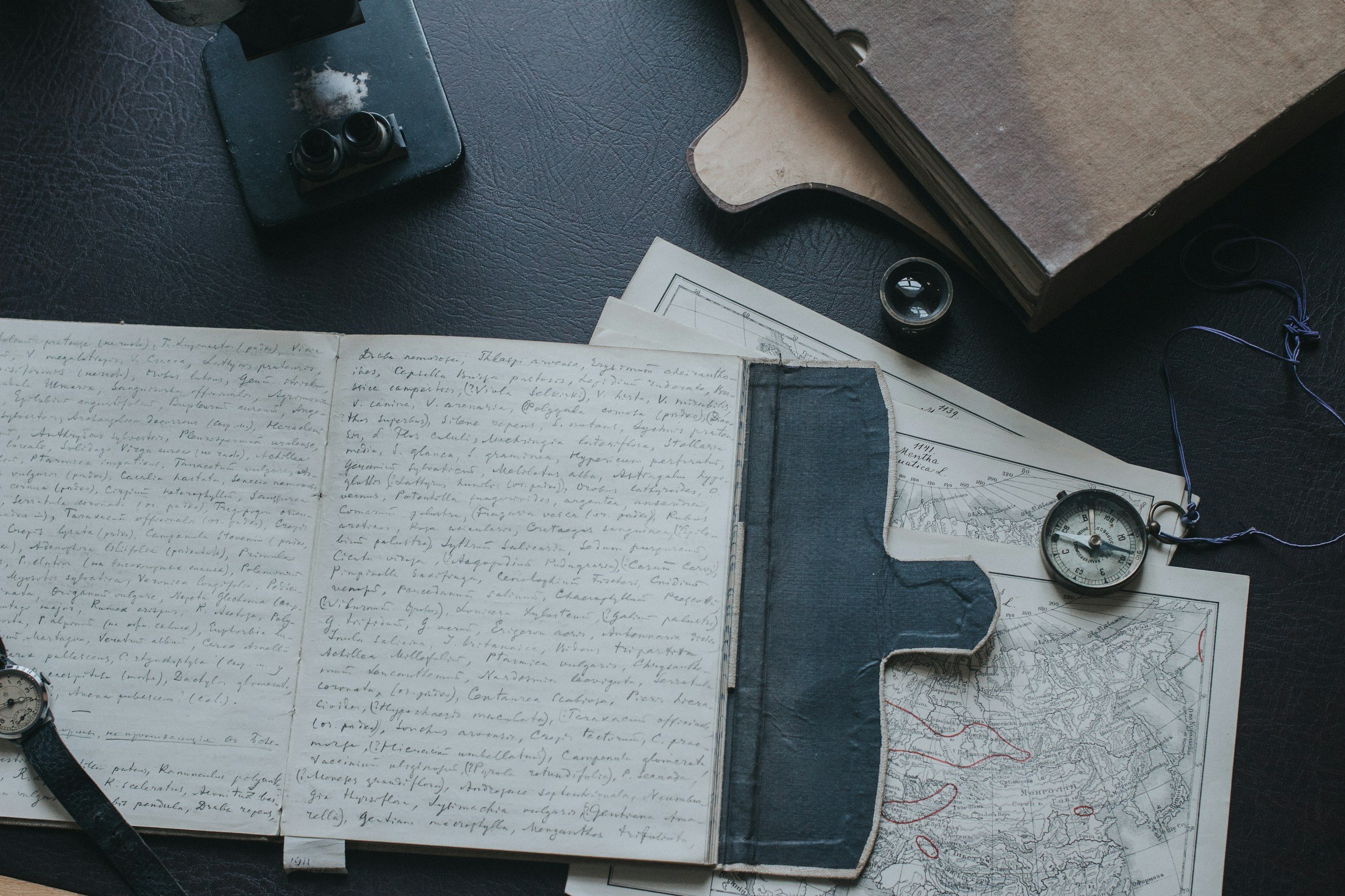 A desk scattered with papers, maps, a compact mirror, a monocle, and a notebook with handwritten notes, indicating a workspace for research or planning.