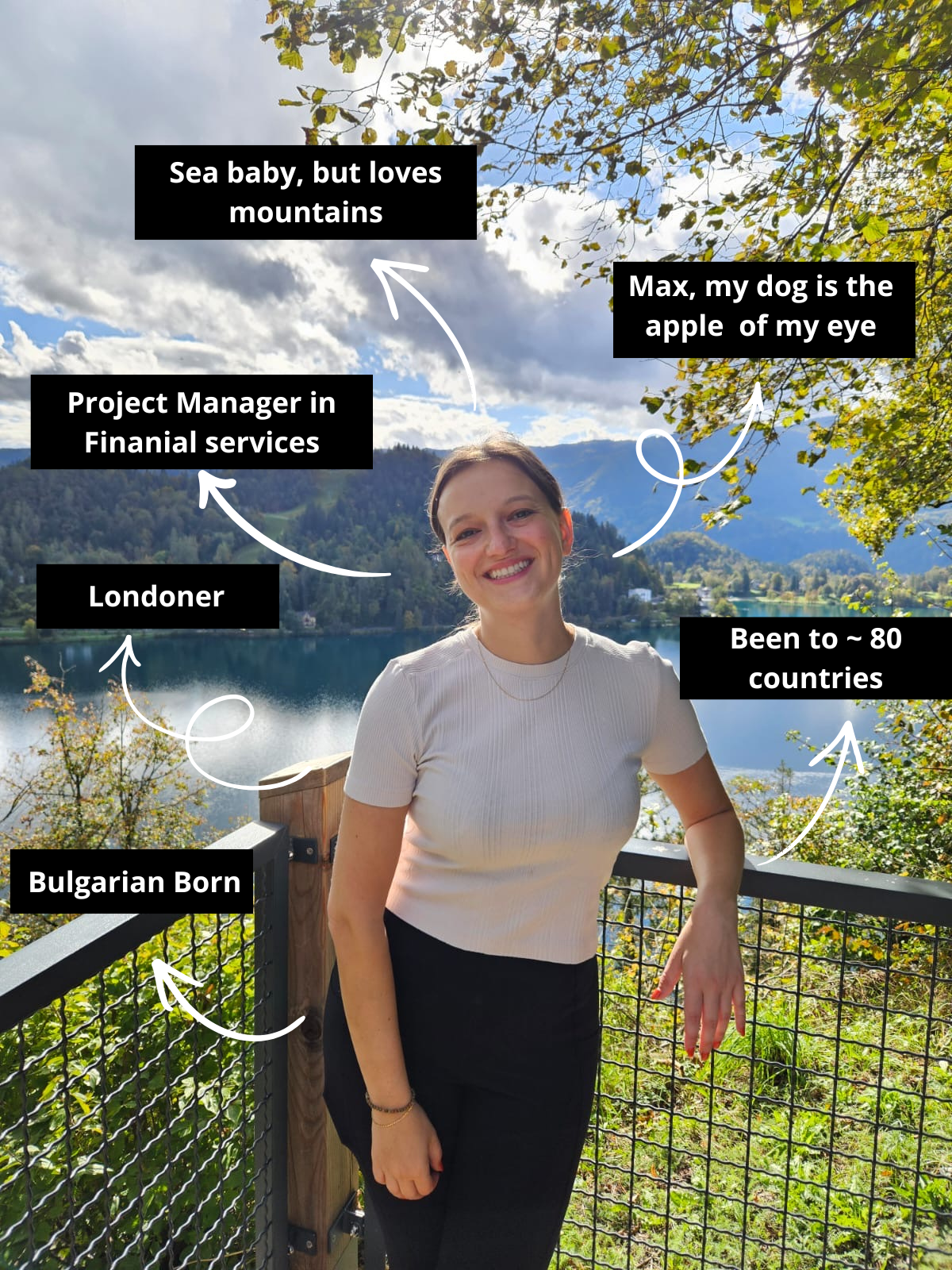 A woman smiling standing outdoors by a lake with mountains in the background, surrounded by trees with green and yellow leaves, during daytime clouds and blue sky.