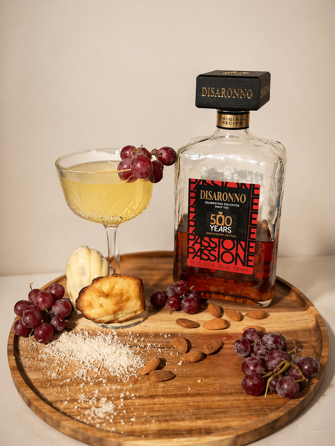 A wooden tray with a bottle of Disaronno Passion liqueur, a cocktail glass with yellow drink garnished with red grapes, a piece of toasted bread, a scoop of ice cream, scattered almonds, shredded coconut, and additional red grapes.