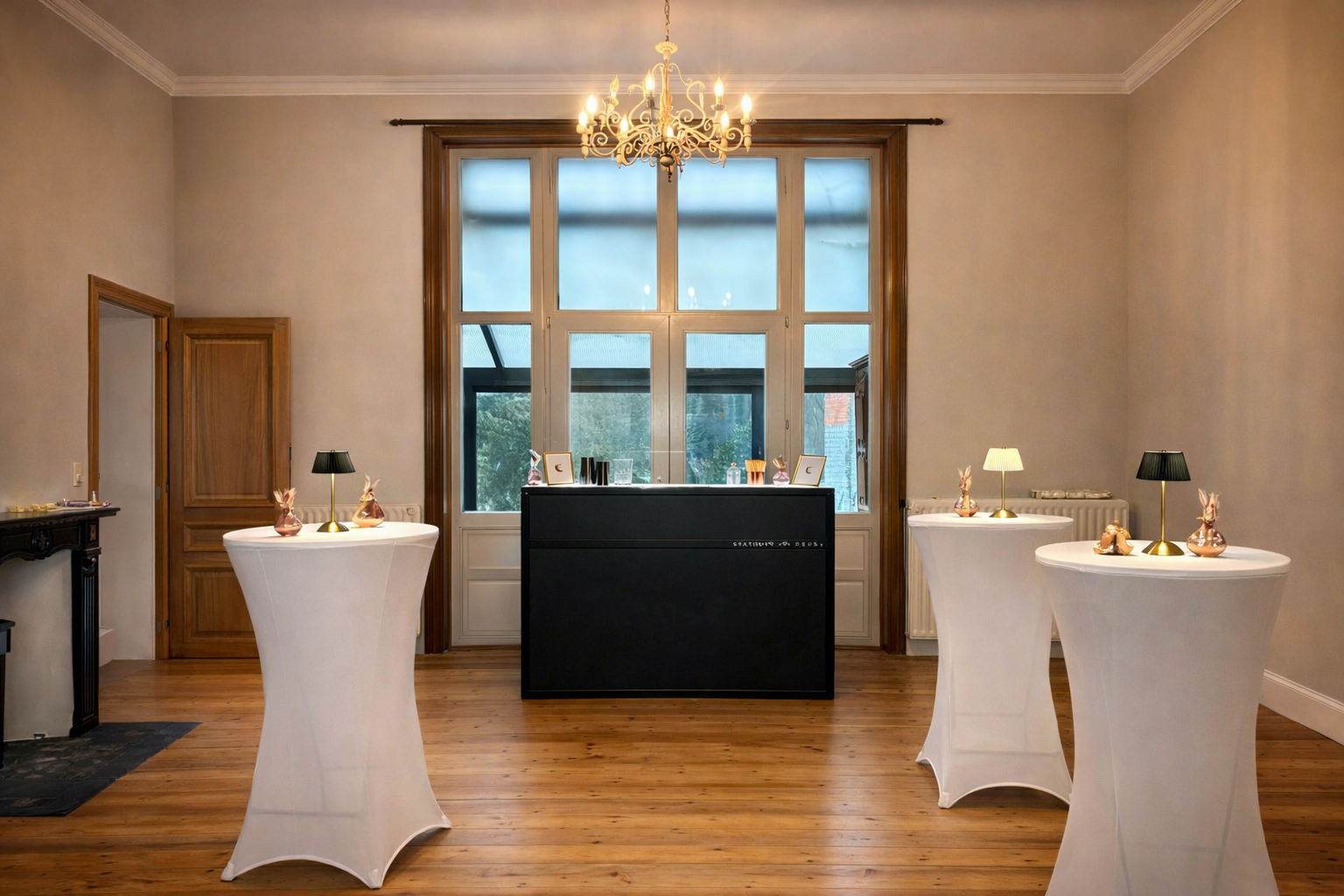 A decorated event room with wooden floors, large window, chandelier, and three high tables with white covers and small lamps and decorative figures on top.