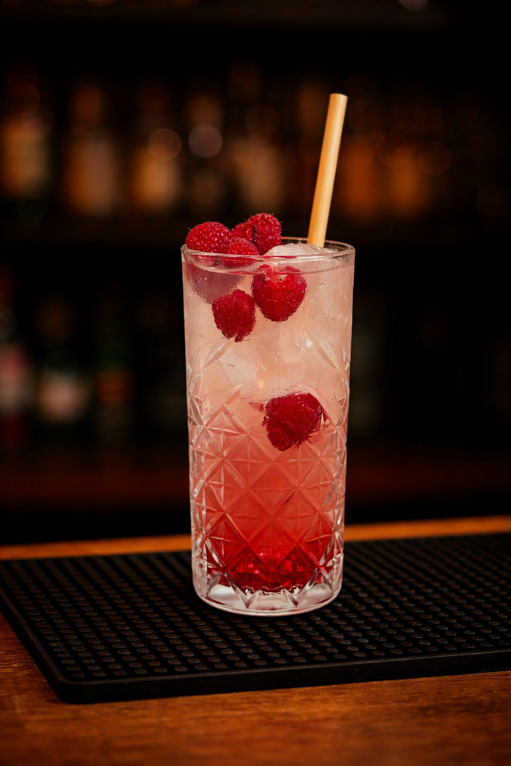 A tall glass cocktail with raspberries and ice, garnished with additional raspberries, and a straw, on a bar counter.