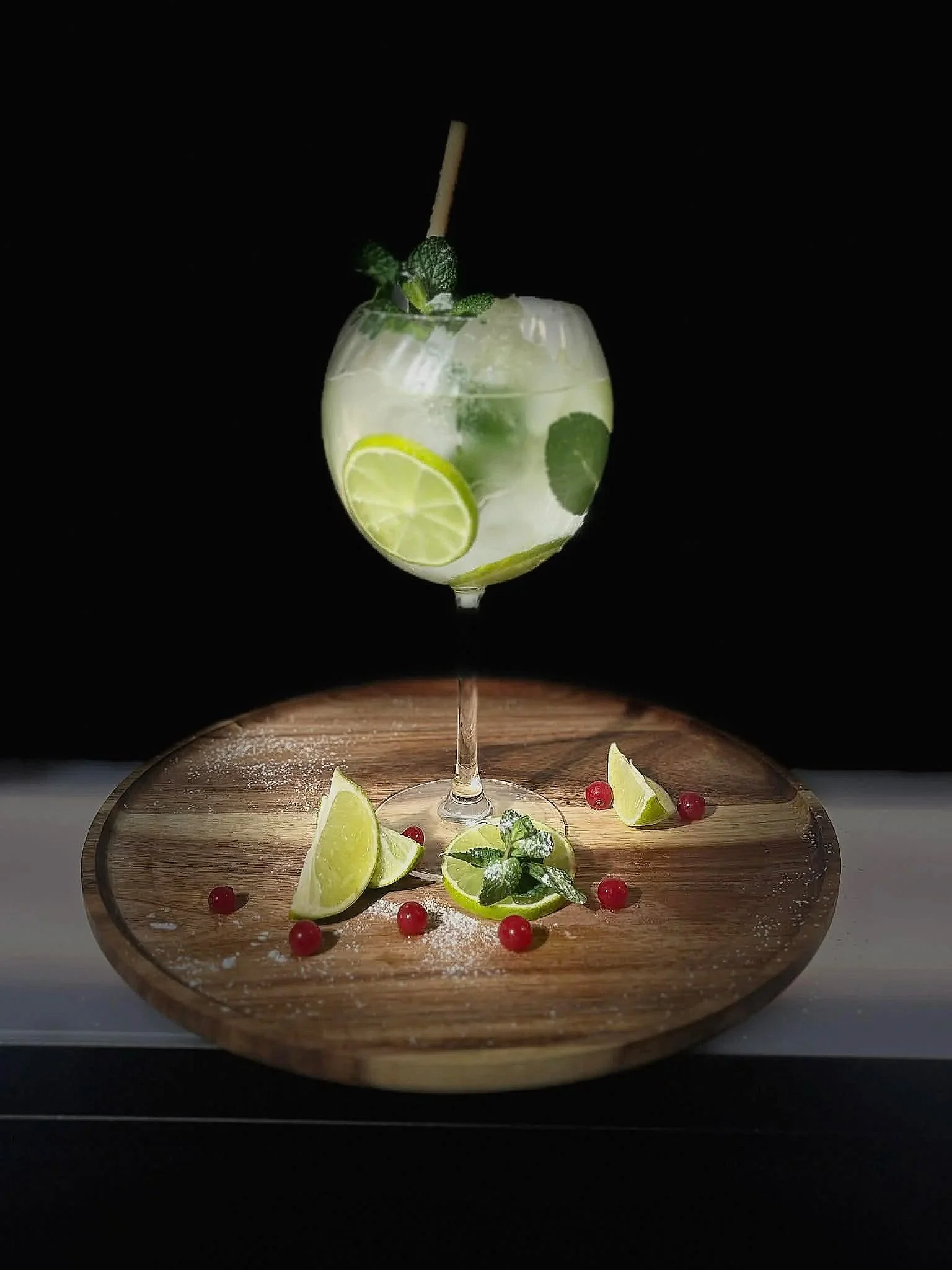 A cocktail glass filled with a clear drink, ice, lemon slices, and mint leaves, with additional garnishes of lime wedges, red berries, and mint sprigs on a wooden board.