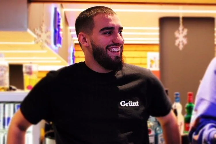 A man with short hair and a beard smiling, wearing a black "Gruit" t-shirt, in a brightly lit indoor setting with festive decorations.