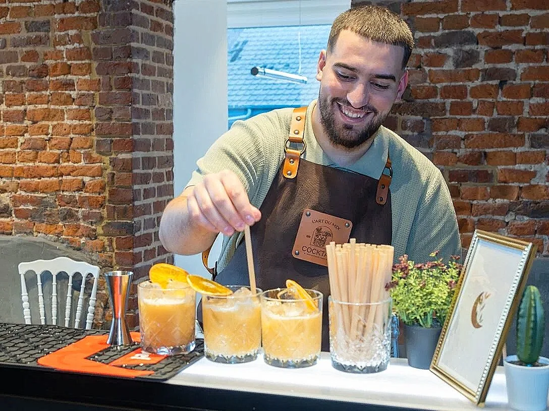 Bartender smiling behind bar preparing four orange cocktails with lemon slices, whisky stones, and straws, surrounded by bar tools, plants, and a framed sign.