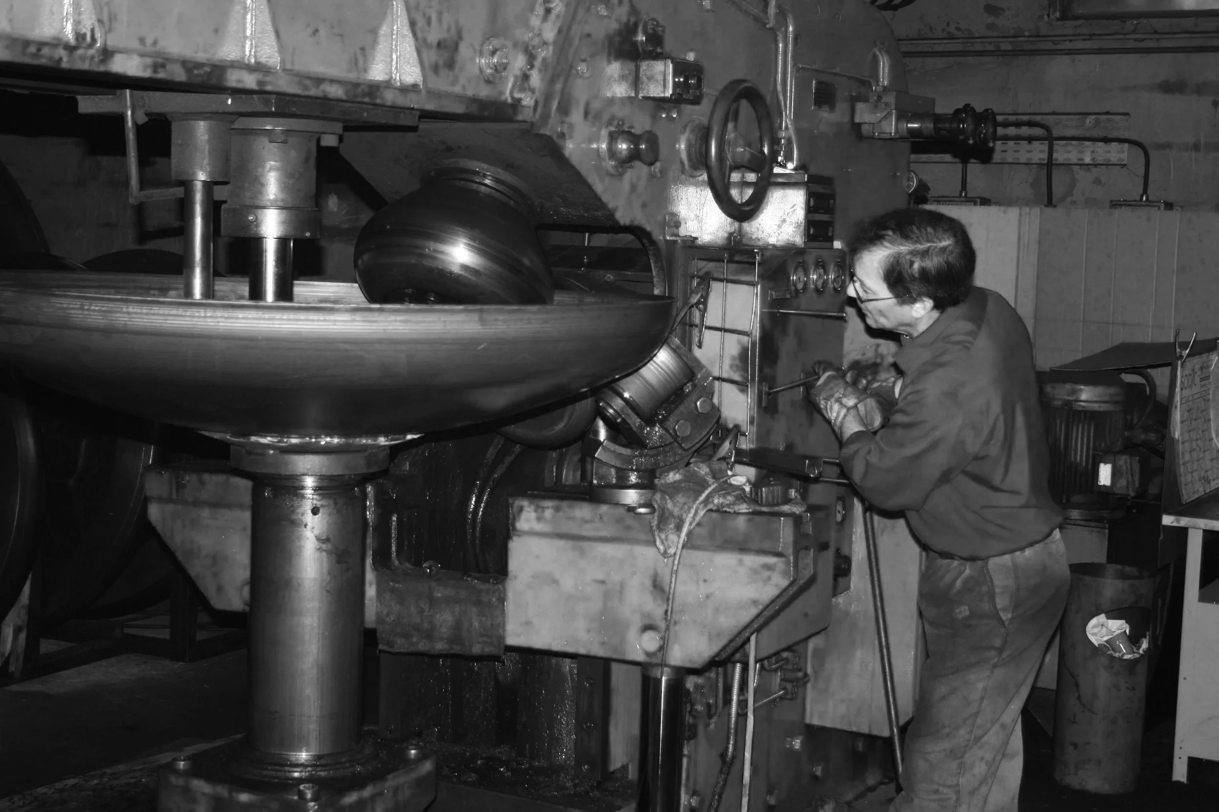 Une personne travaillant sur une machine de grande taille dans un environnement industriel. La machine, une bordeuse, transforme une pièce mécanique complexe en métal, un fond bombé avec plusieurs pièces en rotation ou en mouvement.