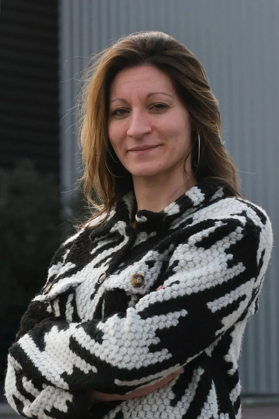 Une femme souriante avec des cheveux bruns, portant un pull à rayures noires et blanches, posant à l'extérieur avec un mur en métal en arrière-plan.