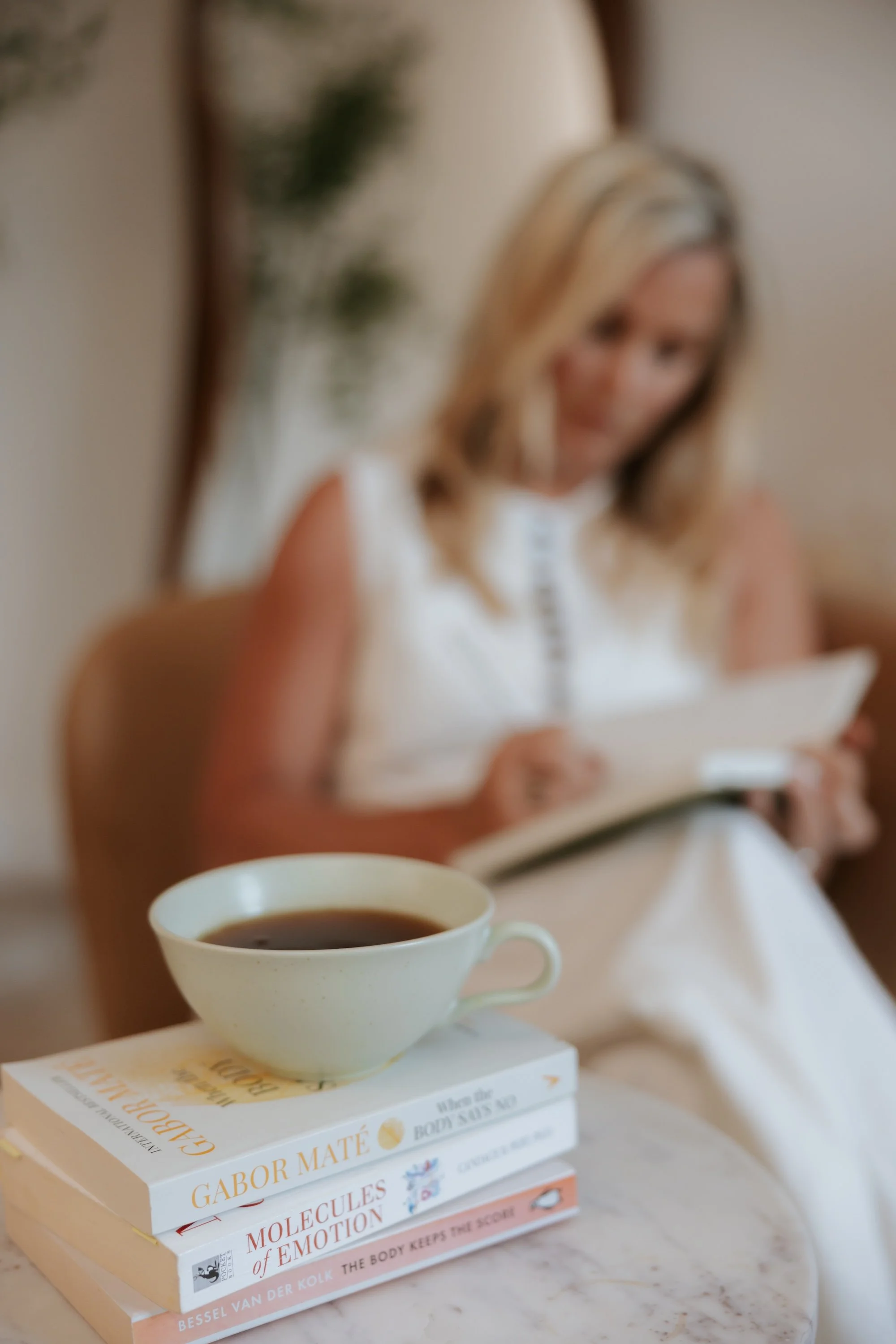 A close-up of a white coffee cup filled with coffee placed on top of a stack of books, with a woman in the background reading a book.
