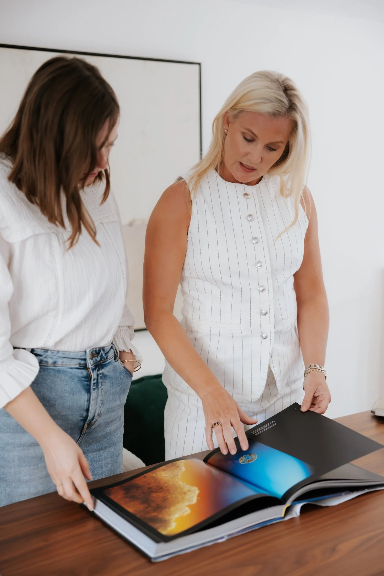 Two women are looking at a large opened book or magazine with colorful images, including a sunset or sunrise landscape, on a wooden table. One woman has blonde hair and wears a white sleeveless top, while the other has brown hair and wears a white blouse and jeans.
