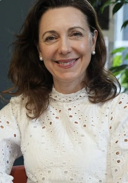 A woman with medium-length brown hair, wearing pearl earrings and a white, eyelet blouse, smiling in an indoor setting with green plants in the background.