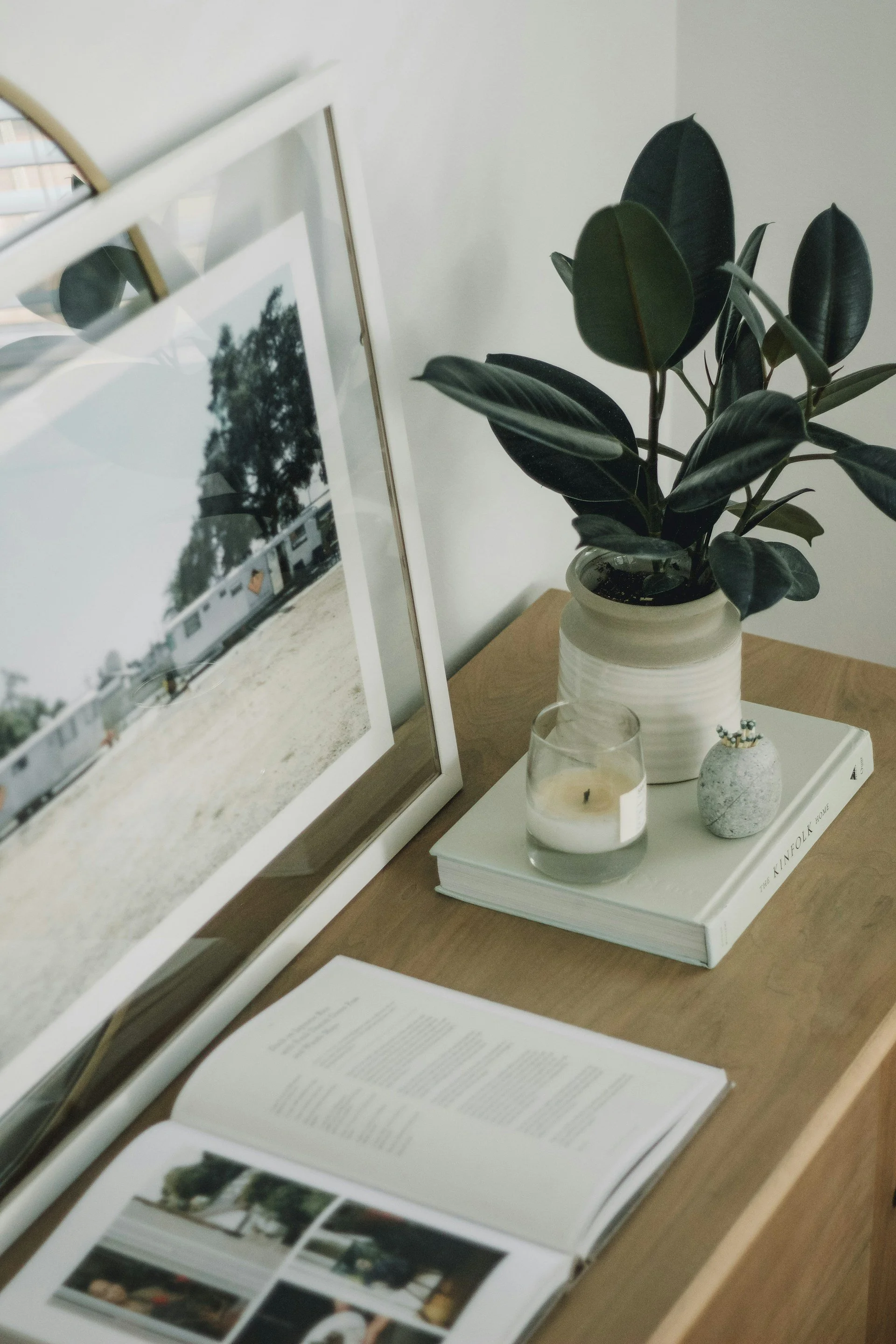A framed photograph leaning on a white surface, a leafy plant in a white ceramic pot, a lit candle in a glass, a small decorative rock with a plant, and a large open book with photographs and text on its pages, all arranged on a wooden table.