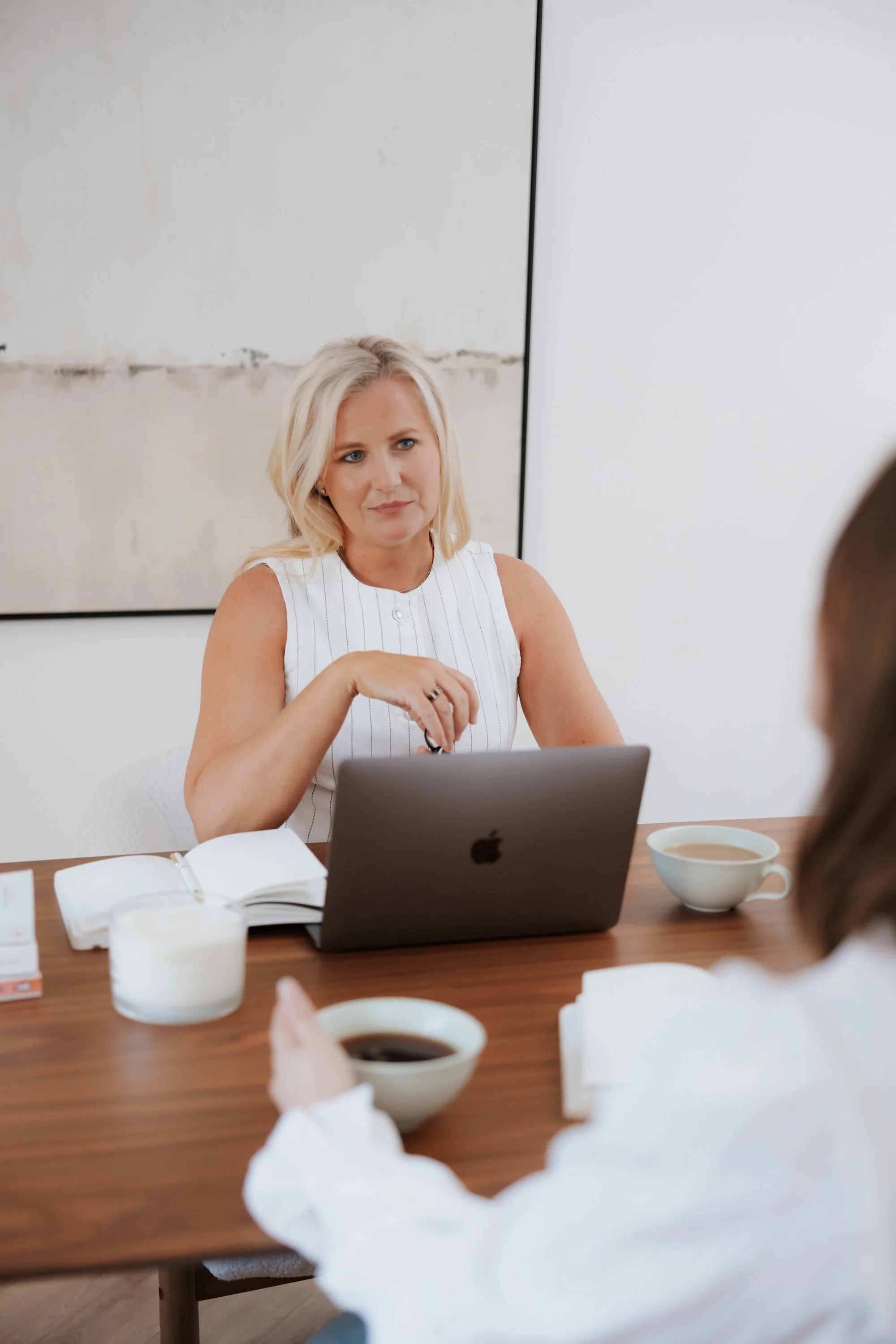 white top is sitting at a wooden table during a meeting, with an open laptop, a notebook, a pen, and a cup of coffee in front of her. She is talking to someone off-camera, with several cups of coffee and tissue boxes on the table, and a plain white wall with a large abstract painting in the background.