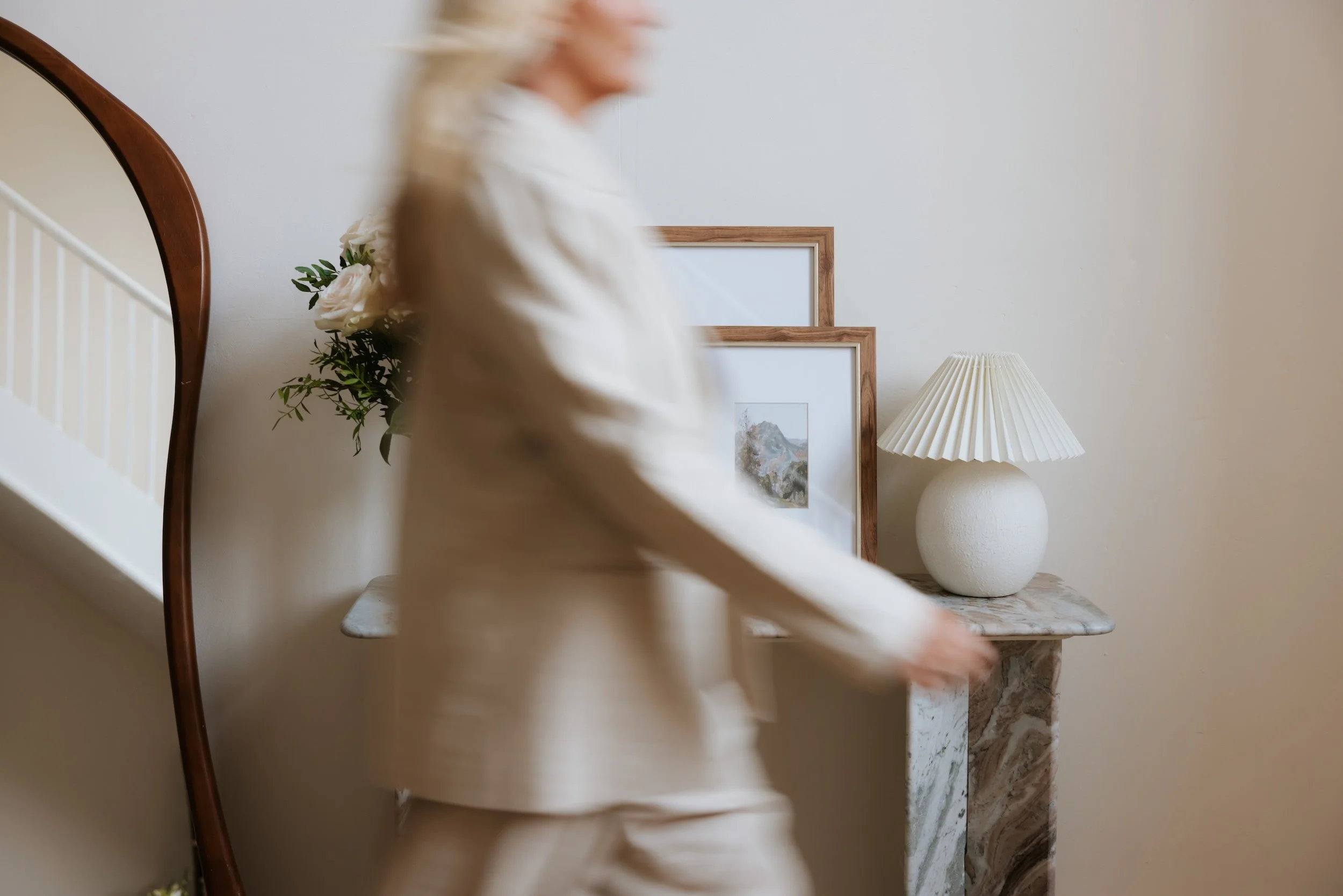 Blurred motion of a blonde woman walking past a side table with a marble top, a decorative lamp, and framed pictures, with a plant in the background.