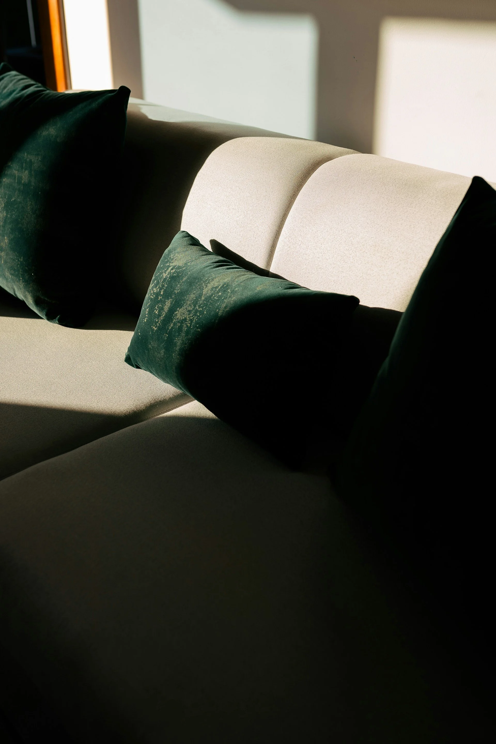 Close-up of a beige sofa with dark green pillows, illuminated by natural sunlight, creating shadows on the wall.
