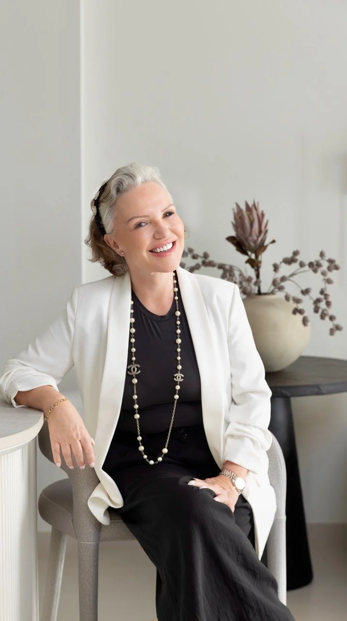 A smiling elderly woman with gray hair, dressed in a white blazer and black outfit, sitting on a chair in a room with a beige wall and a decorative vase with plants in the background.