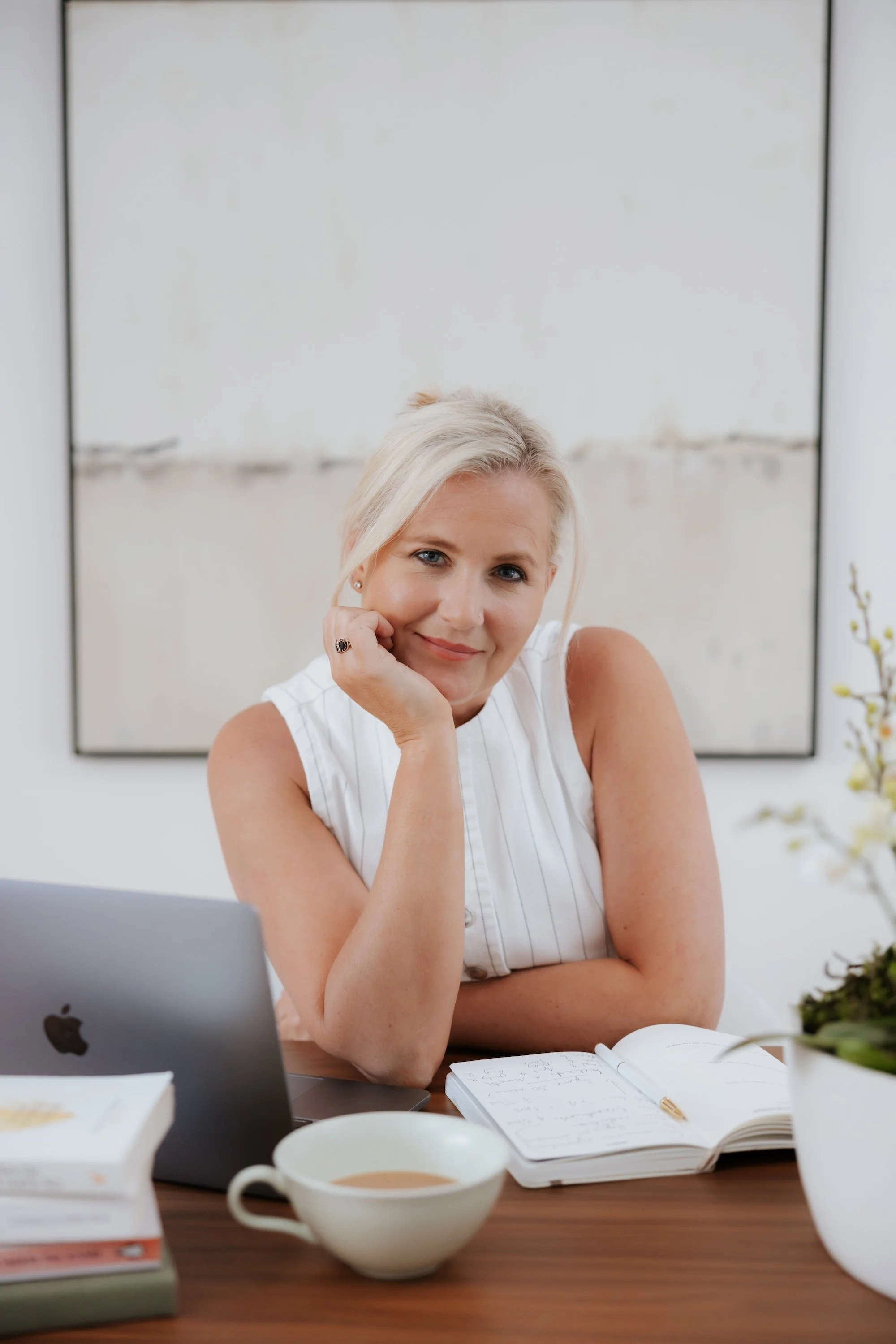 A woman with blonde hair sits at a desk, resting her chin on her hand, smiling. She has a laptop, a notebook with handwritten notes, a gold pen, a cup of coffee, and a stack of books on the desk. In the background, there's a large mirror and some plants.