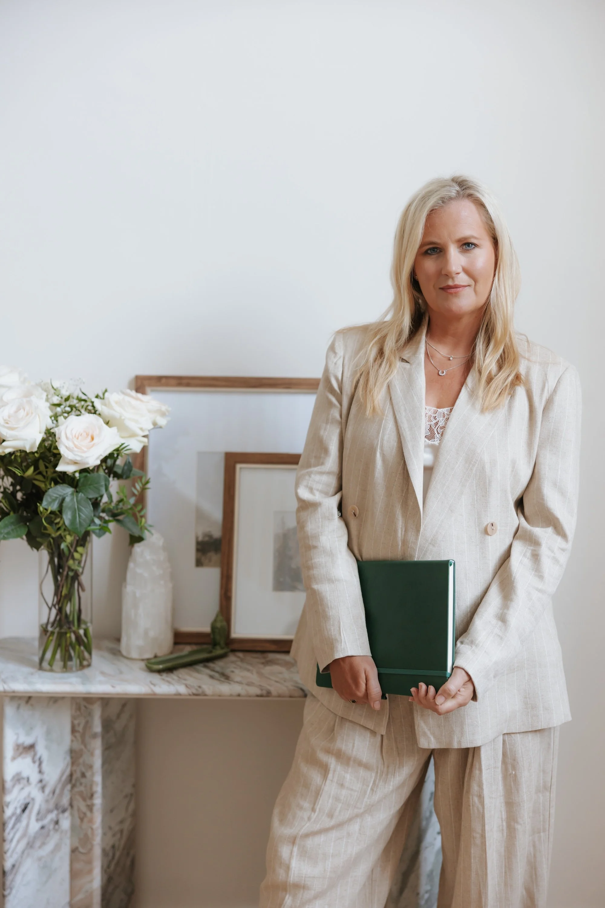 Catherine Raincock fertility specialist dressed in a beige plaid suit holding a green file folder stands in a room with a white wall. There are white roses in a glass vase and framed pictures on a marble console in the background.
