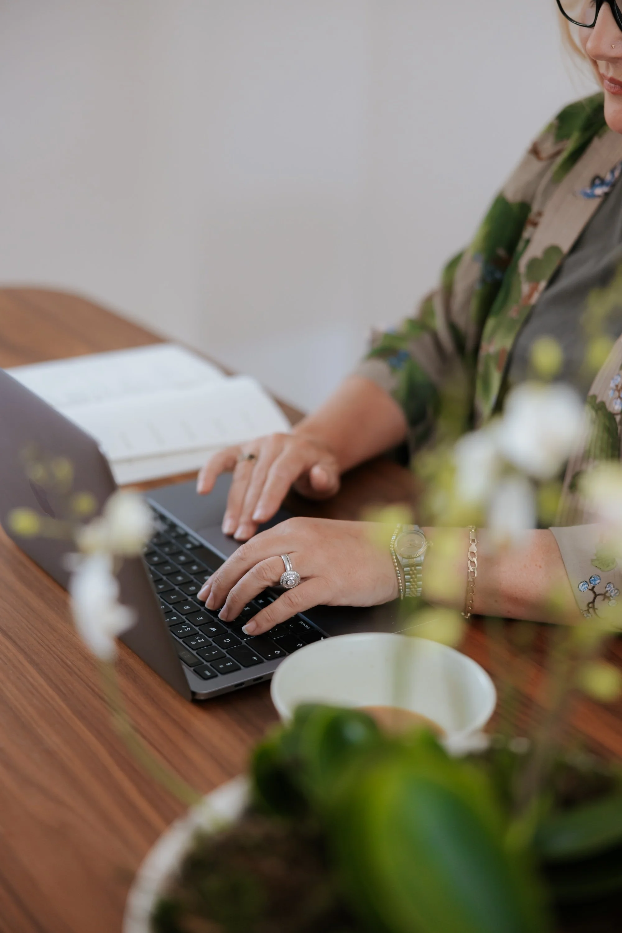 Close-up of a woman typing on a laptop at a wooden table, with a white cup, a floral plant in the foreground, and a plain background.