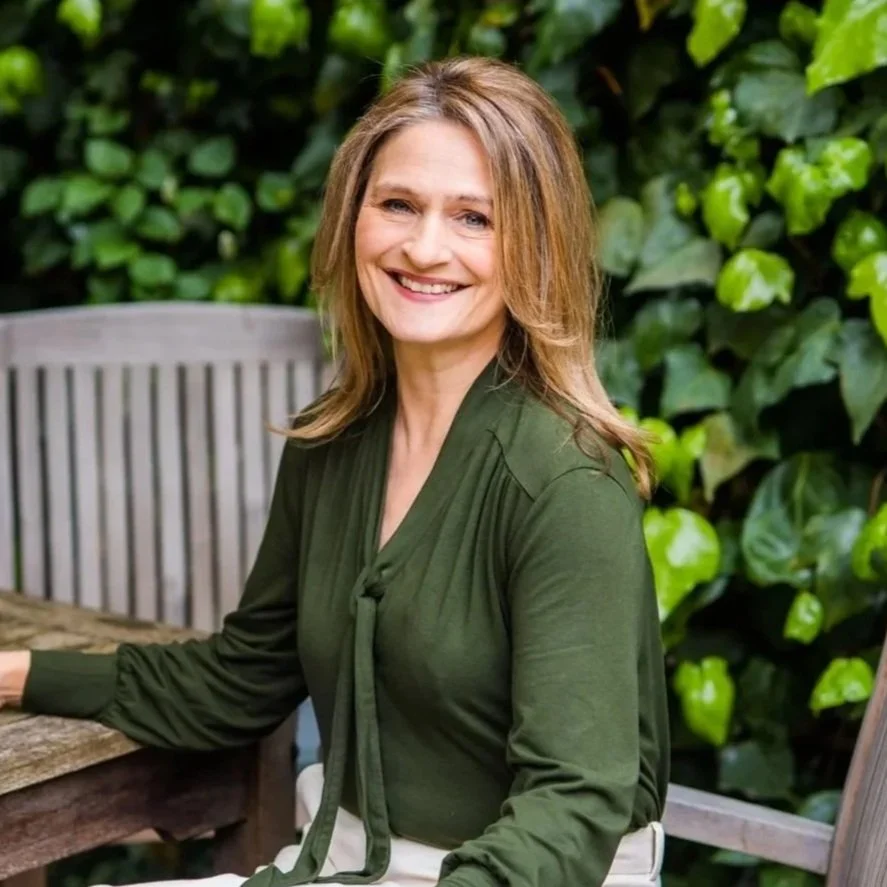 A woman with shoulder-length brown hair, smiling, wearing a green blouse, sitting outdoors at a wooden table with lush green foliage in the background.