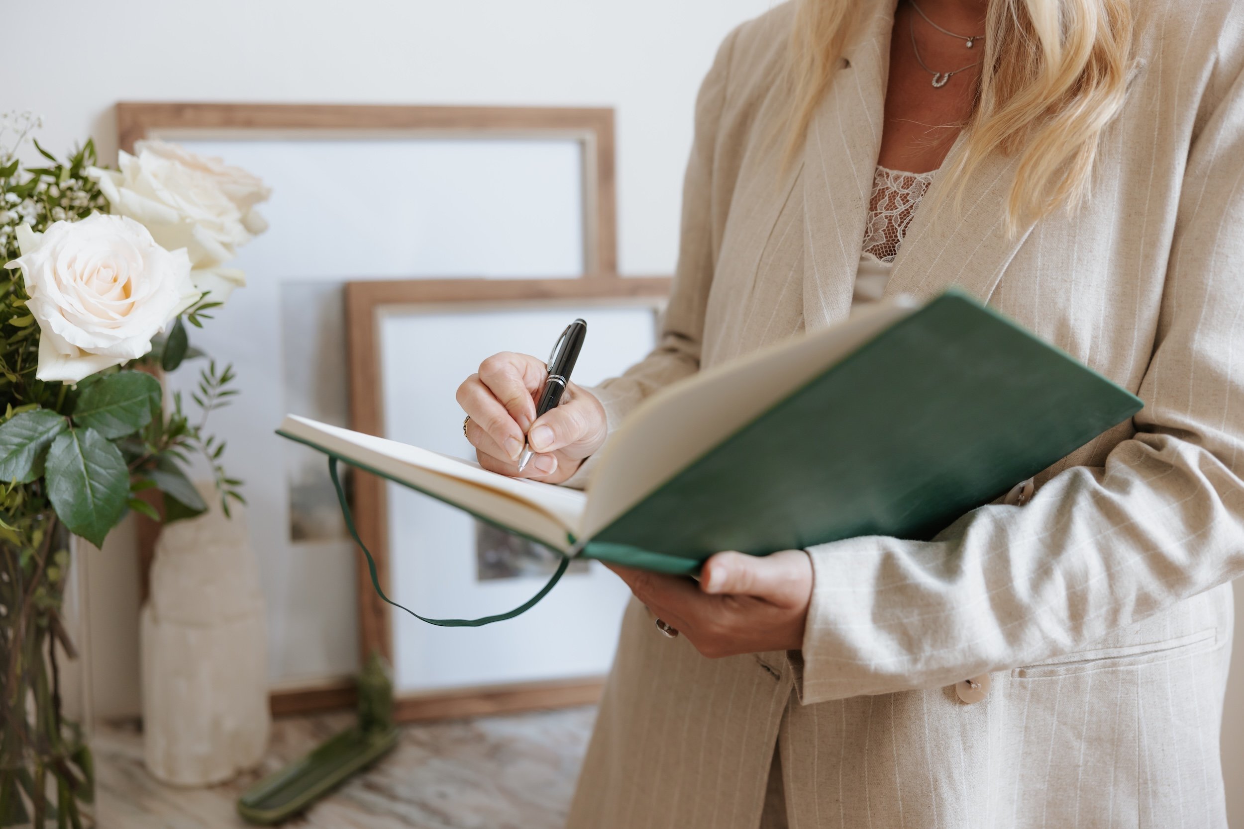 A woman in a beige pinstripe suit holding a green notebook and writing with a pen. In the background, there are framed pictures and a vase with white roses and greenery.